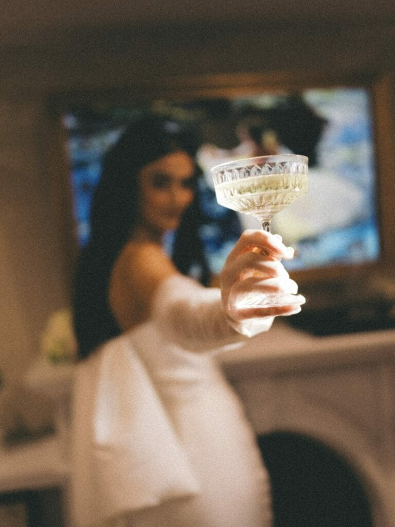A woman in a white dress holds a glass of champagne toward the camera, her face blurred in the background.
