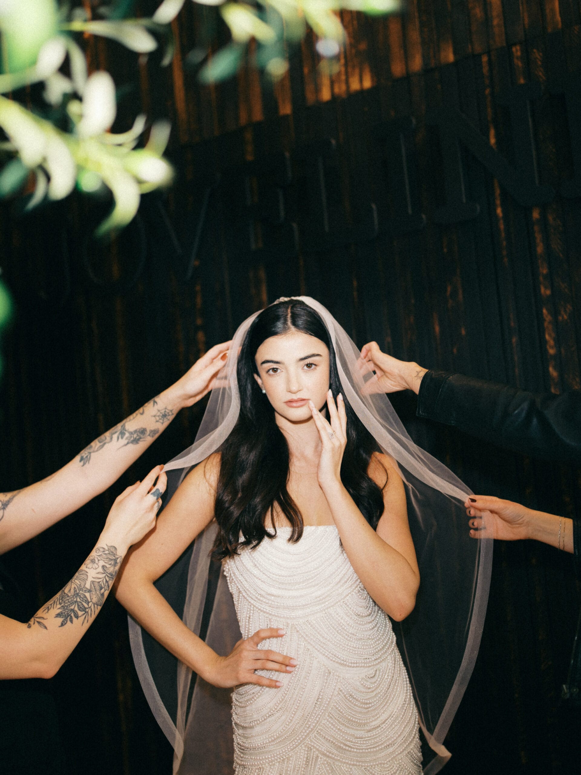 A bride in a beaded dress has her veil adjusted by three people, standing against a dark wooden background.