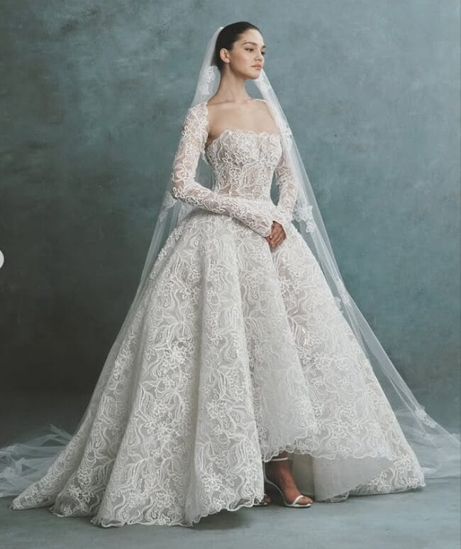 A bride in an elegant lace wedding gown with long sleeves and a long veil stands against a gray backdrop, ready to inspire at upcoming Bridal Trunk Shows.